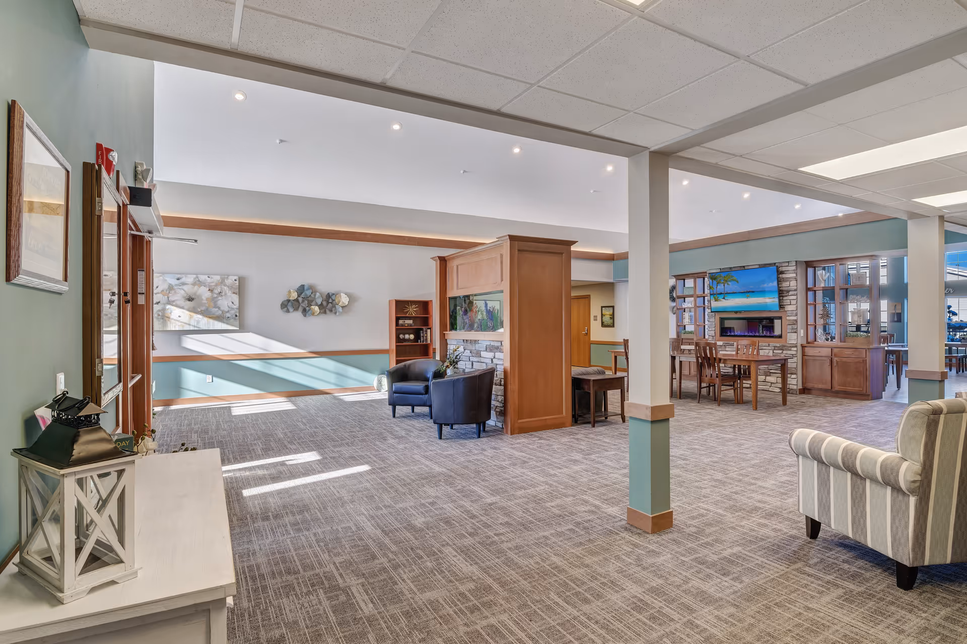 A spacious and well-lit common area in a senior living facility featuring a central wooden divider with a built-in aquarium and stone fireplace. The room has carpeted floors, light blue and beige walls, and recessed ceiling lights. There are several seating areas including black armchairs and a striped armchair. A large flat-screen TV is mounted on a stone wall with wooden shelving units on either side. The space appears inviting and comfortable for residents to relax and socialize.