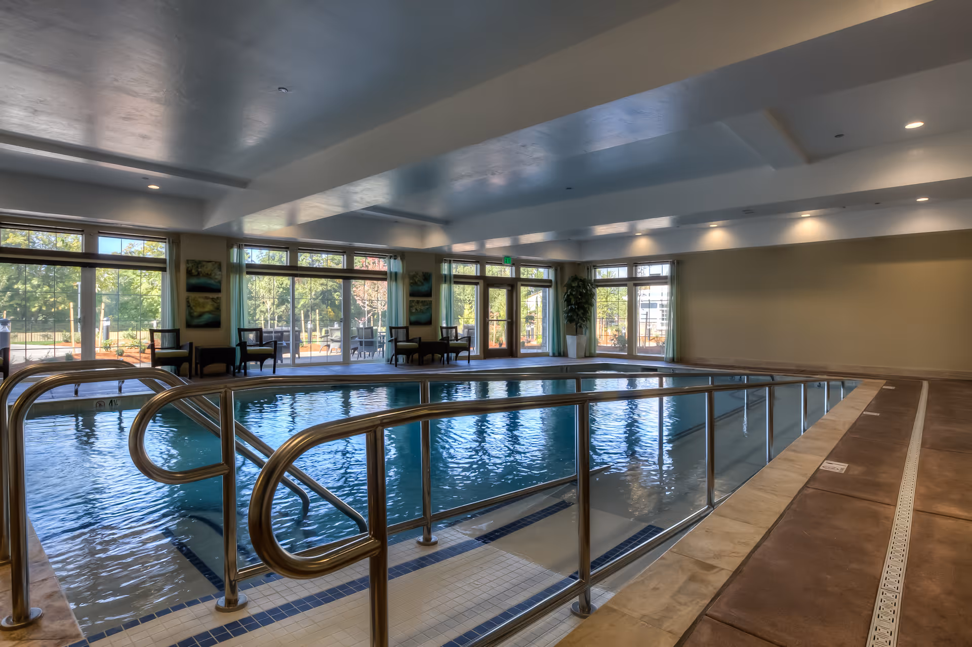 Indoor swimming pool with stainless steel handrails, seating and large windows overlooking landscaped grounds.