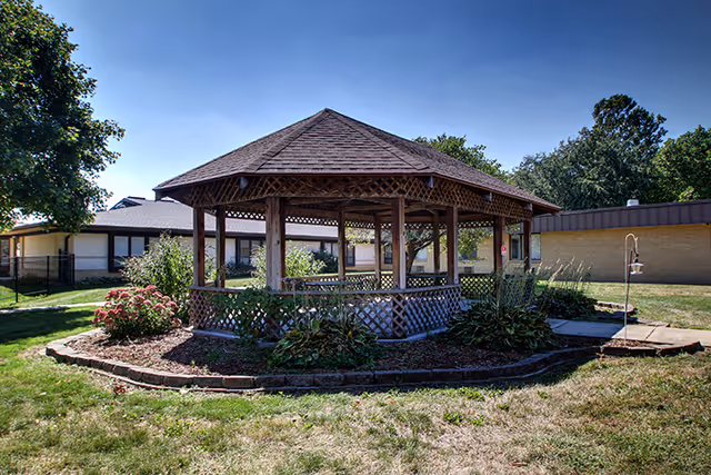 A hexagonal wooden gazebo sits in a landscaped courtyard with grass and single-story buildings in the background.