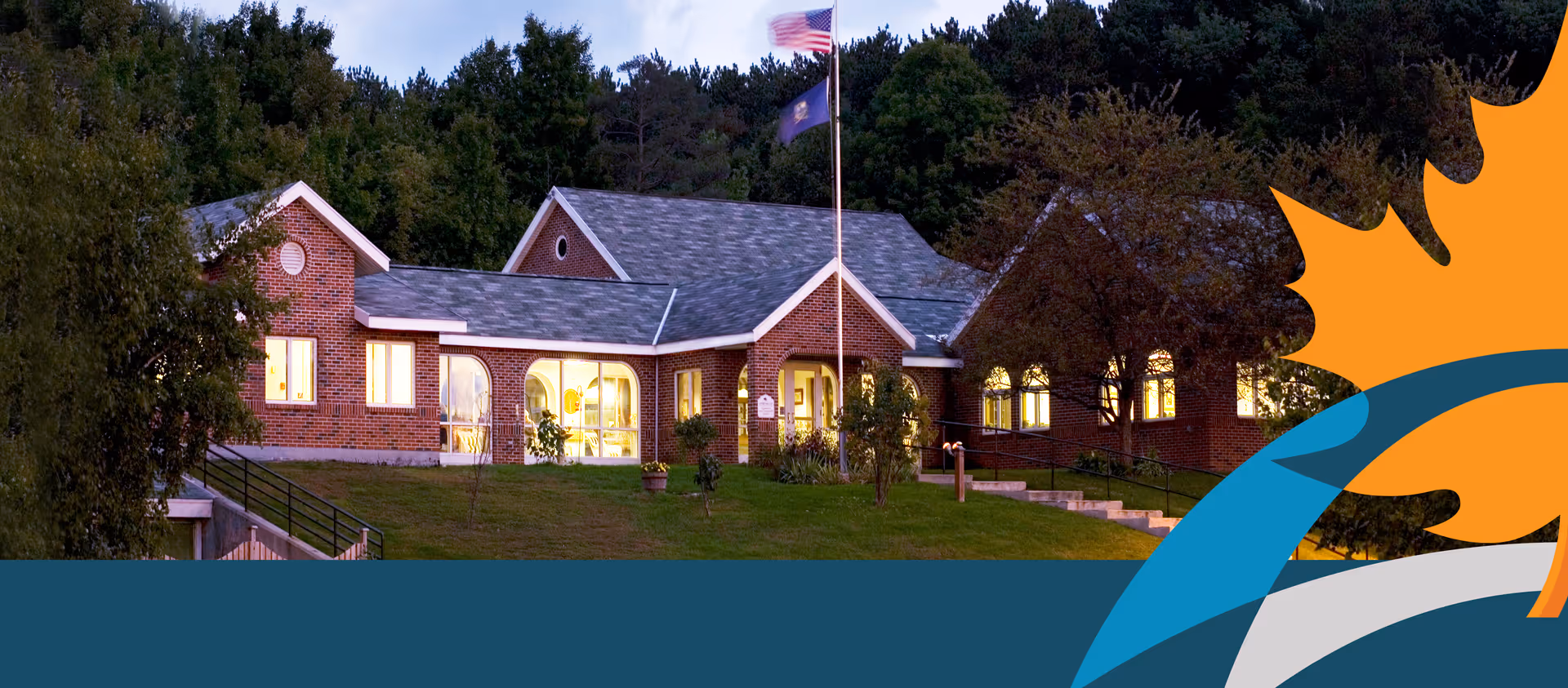 Exterior view of a brick building with large windows lit from inside, surrounded by trees and greenery. An American flag and another flag are flying on a flagpole in front of the building. The sky is dim, indicating early evening or dusk.