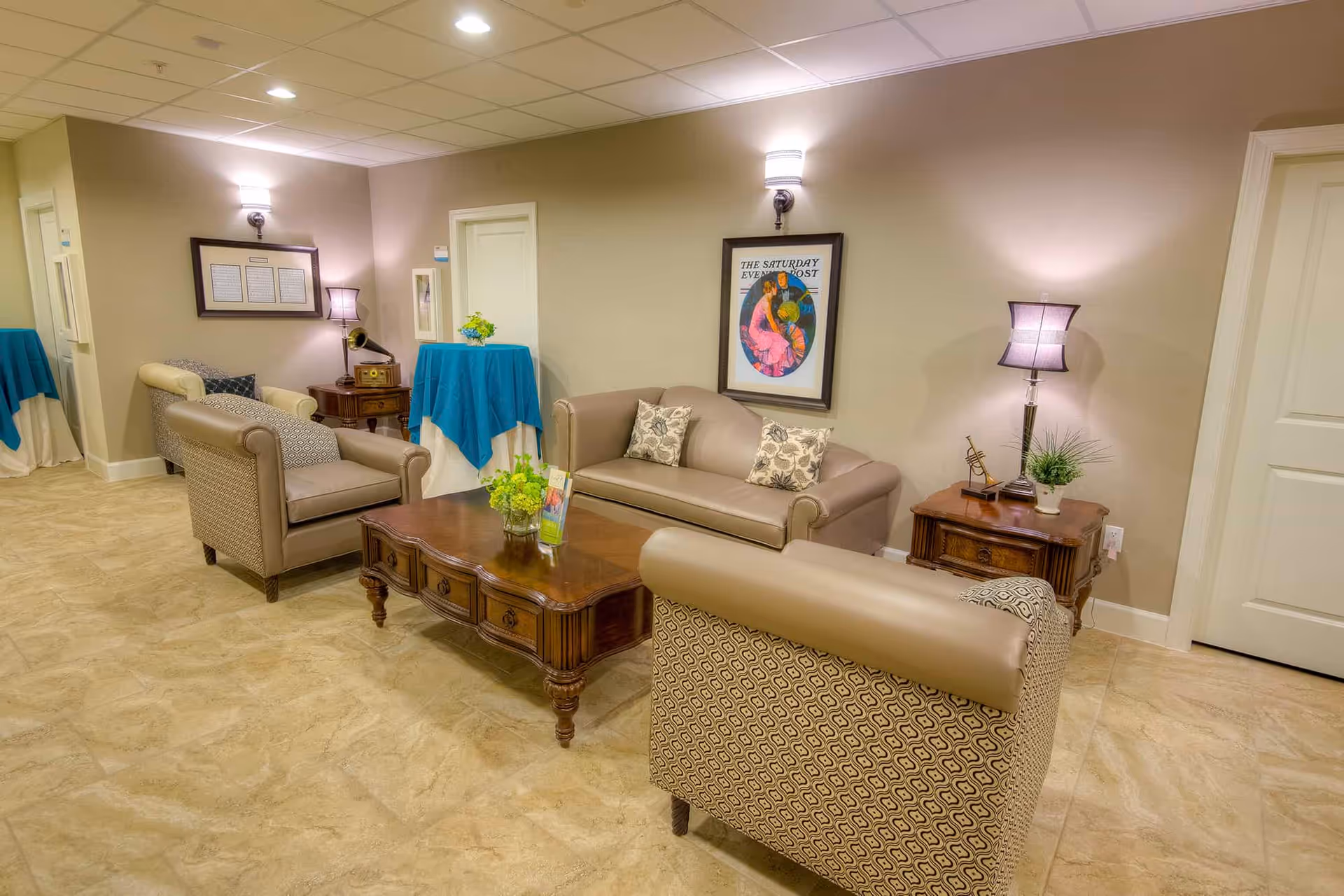Communal sitting area with a sofa and armchairs arranged around a wooden coffee table, side tables, lamps, and framed artwork.