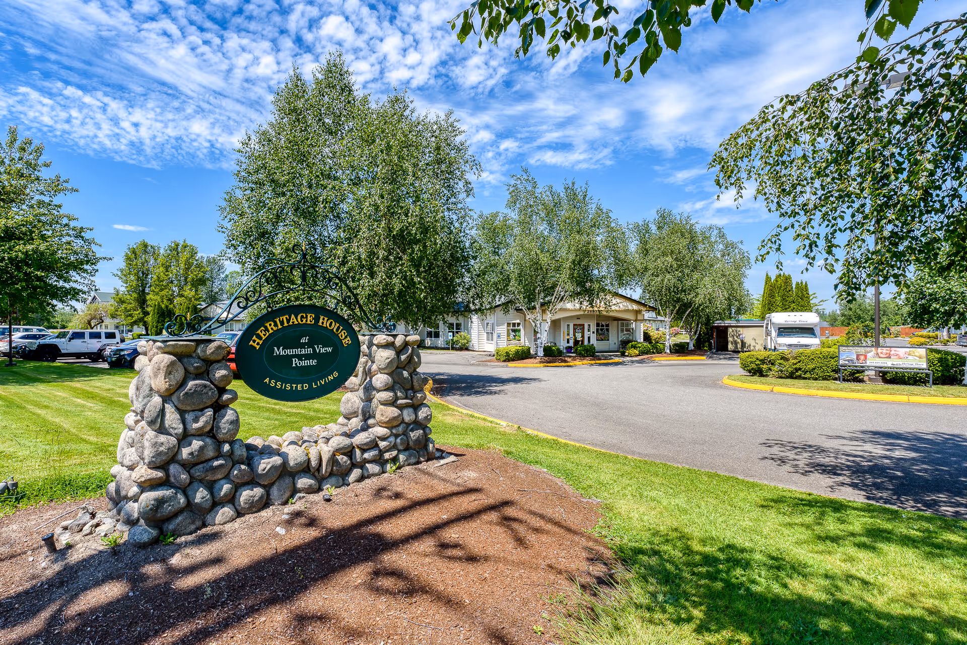 Outdoor view of Heritage House at Mountain View Pointe Assisted Living facility entrance with a stone sign, green lawn, trees, parked cars, and a clear blue sky with scattered clouds.