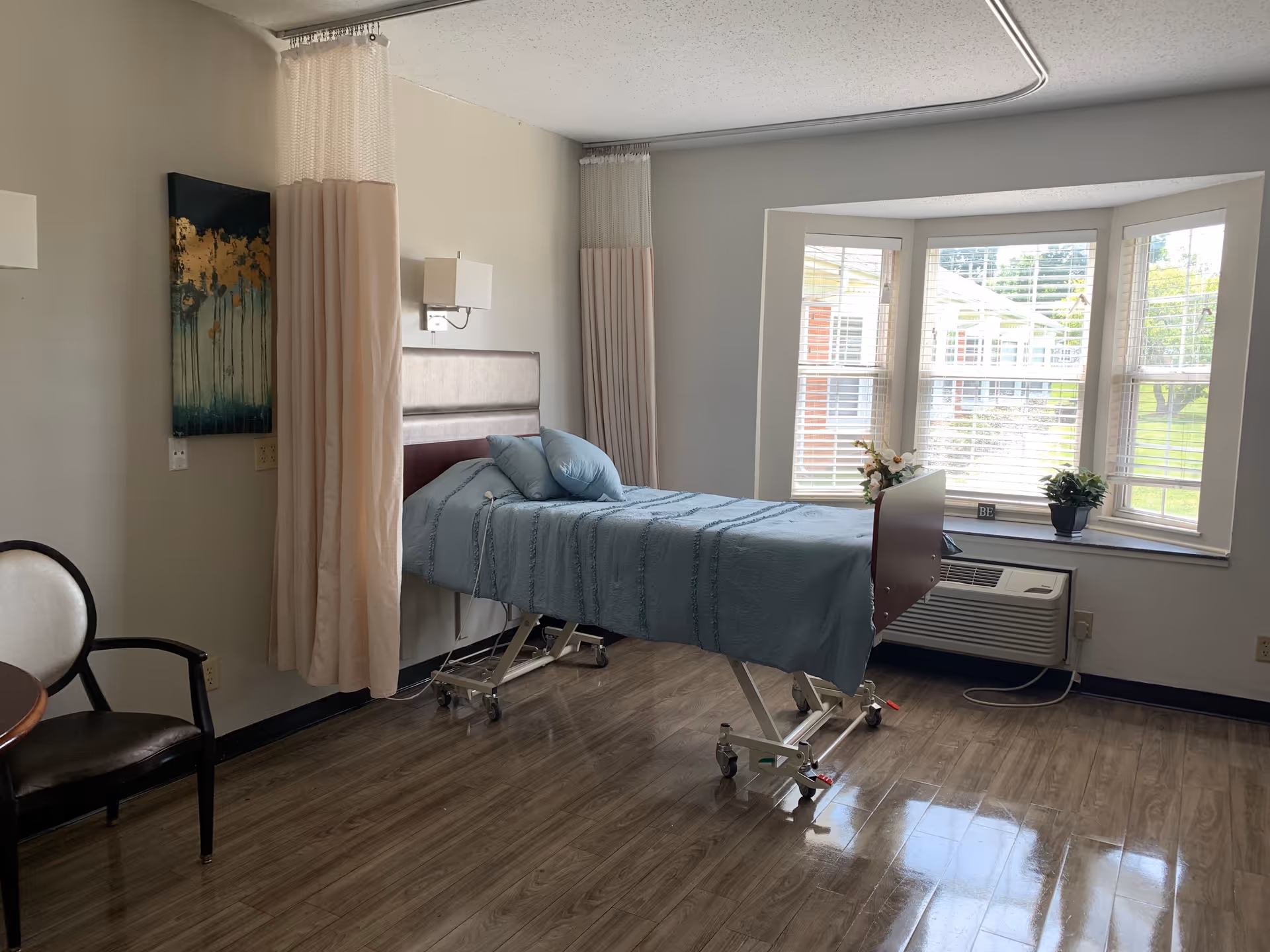 A nursing facility room with a hospital bed covered in a blue blanket and two pillows. The bed is positioned near a large bay window with white blinds, letting in natural light. There are privacy curtains hanging from the ceiling around the bed. A chair with a dark seat and light backrest is placed near a small round table. The floor is wood-patterned, and a wall-mounted light fixture is above the bed. A small air conditioning unit is installed below the window, and there are two small potted plants on the window sill.