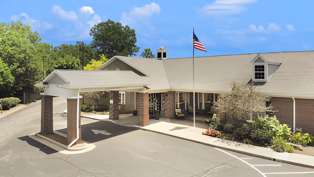 Front exterior view of a senior living facility building with a covered entrance, an American flag on a flagpole, surrounded by greenery and trees under a blue sky with some clouds.