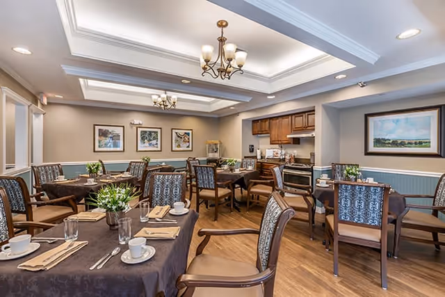 A well-lit dining room with several tables covered in dark tablecloths, each set with cups, glasses, napkins, and silverware. The room features wooden chairs with patterned upholstery, a small kitchen area with wooden cabinets and appliances, framed artwork on the walls, and ceiling light fixtures.
