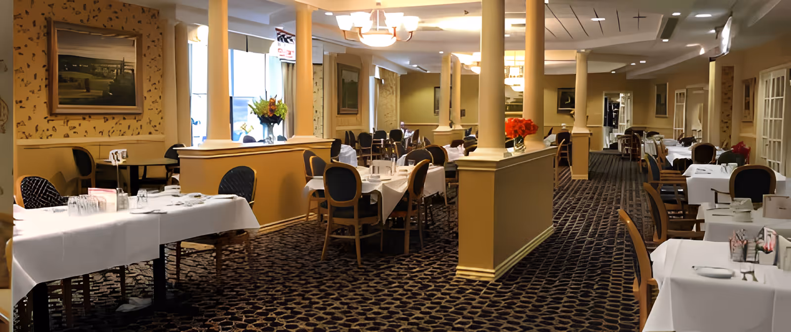 Interior view of a dining room in a senior living facility with tables covered in white tablecloths, chairs arranged around them, decorative columns, floral centerpieces, framed artwork on the walls, and carpeted flooring.