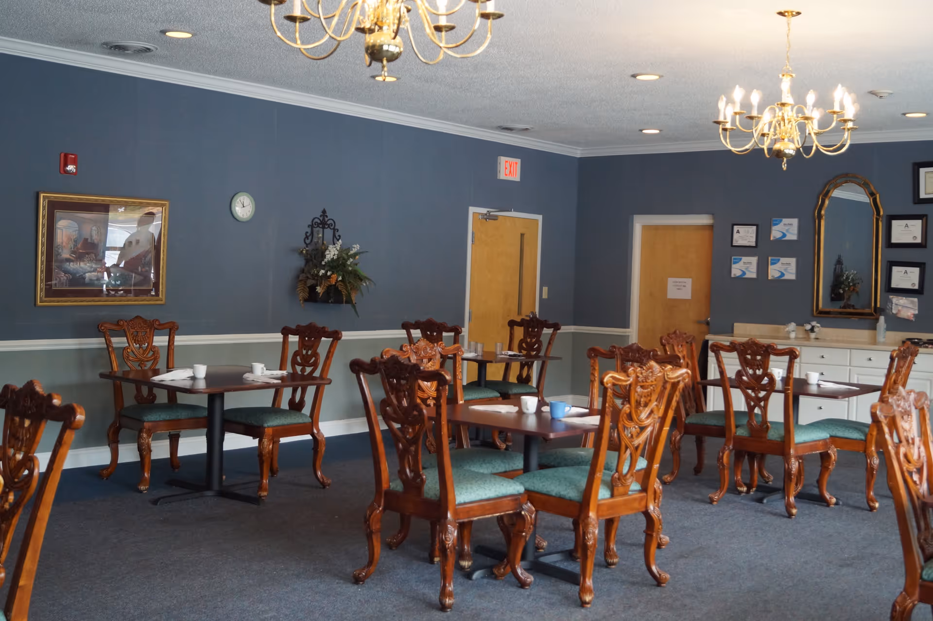 A dining room with several wooden tables and chairs with green cushions. The walls are painted blue with white trim, decorated with a framed picture, a clock, a floral wall arrangement, and a large mirror. There are two chandeliers hanging from the ceiling and two wooden doors, one with an exit sign above it.