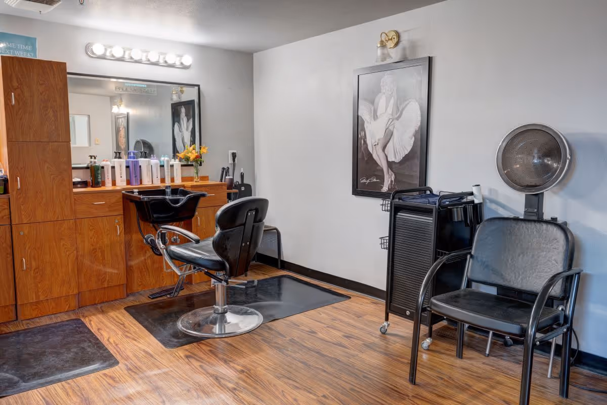Interior view of a salon area with a black salon chair in front of a wooden cabinet and mirror. Various hair care products are lined up on the counter. To the right, there is a black chair with a hair dryer hood attached to the wall above it. A framed black and white picture of Marilyn Monroe is hanging on the wall.
