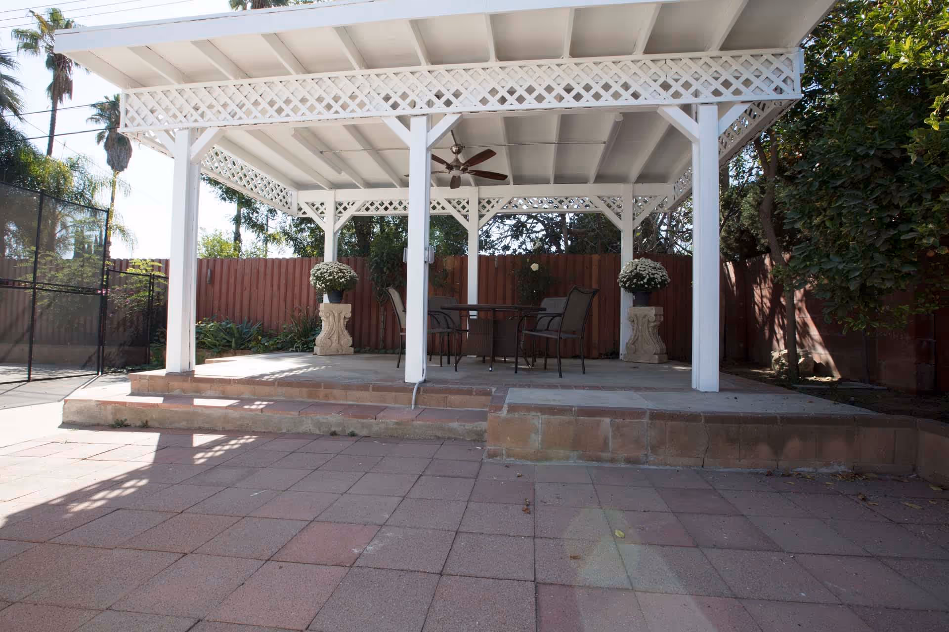 Covered outdoor patio with a white lattice pergola, table and chairs, ceiling fan and potted plants.
