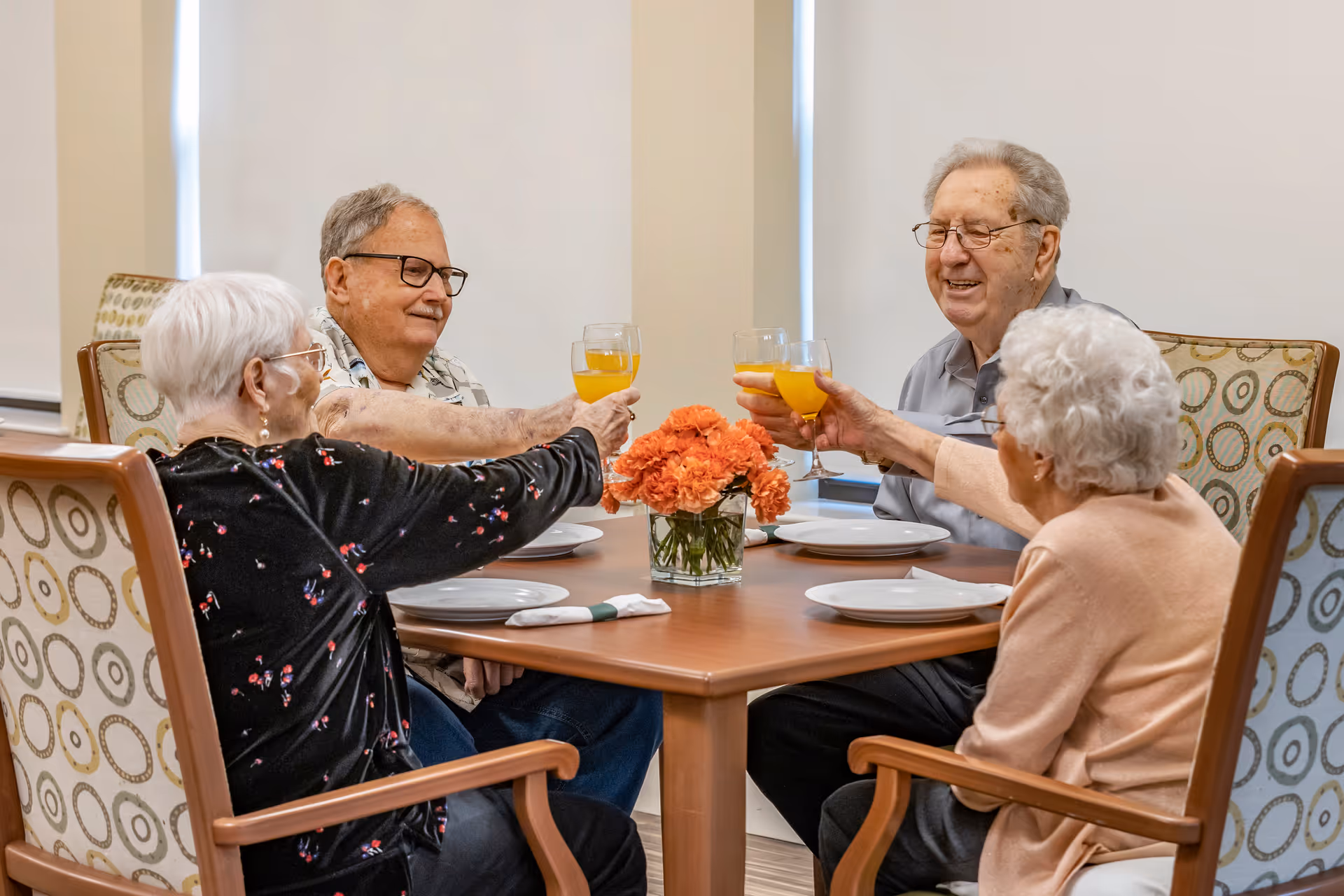 Four elderly people sitting around a wooden dining table, raising glasses of orange juice in a toast. The table is set with white plates, napkins, and a vase of orange flowers in the center. The chairs have patterned upholstery, and the background shows closed window blinds.