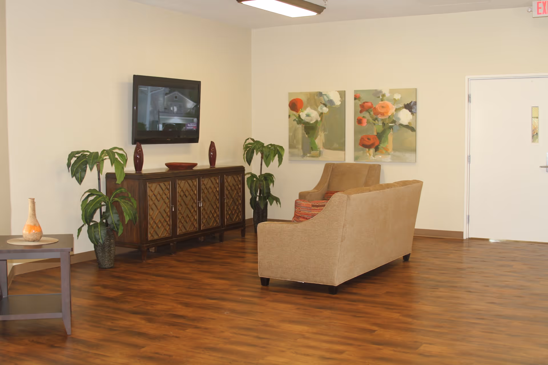 A clean common room with a sofa facing a wall-mounted TV above a wooden cabinet, potted plants, and floral artwork.