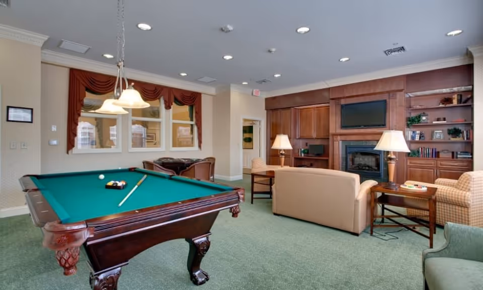 A cozy recreational room featuring a green felt pool table with balls and a cue stick, a beige sofa, armchairs, wooden side tables with lamps, a built-in wooden bookshelf, and a fireplace with a mounted flat-screen TV above it. The room has green carpet flooring, cream-colored walls, and windows with red valances.