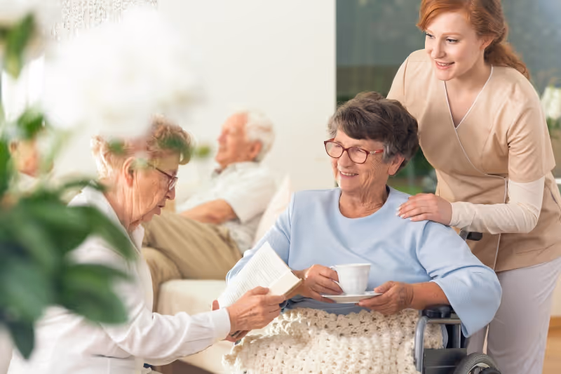 A smiling elderly woman in a wheelchair holding a cup and saucer, accompanied by a caregiver who is gently touching her shoulder. Another elderly woman is sitting nearby reading a book, and an elderly man is seated in the background. The setting appears to be a bright and comfortable indoor common area with plants visible.