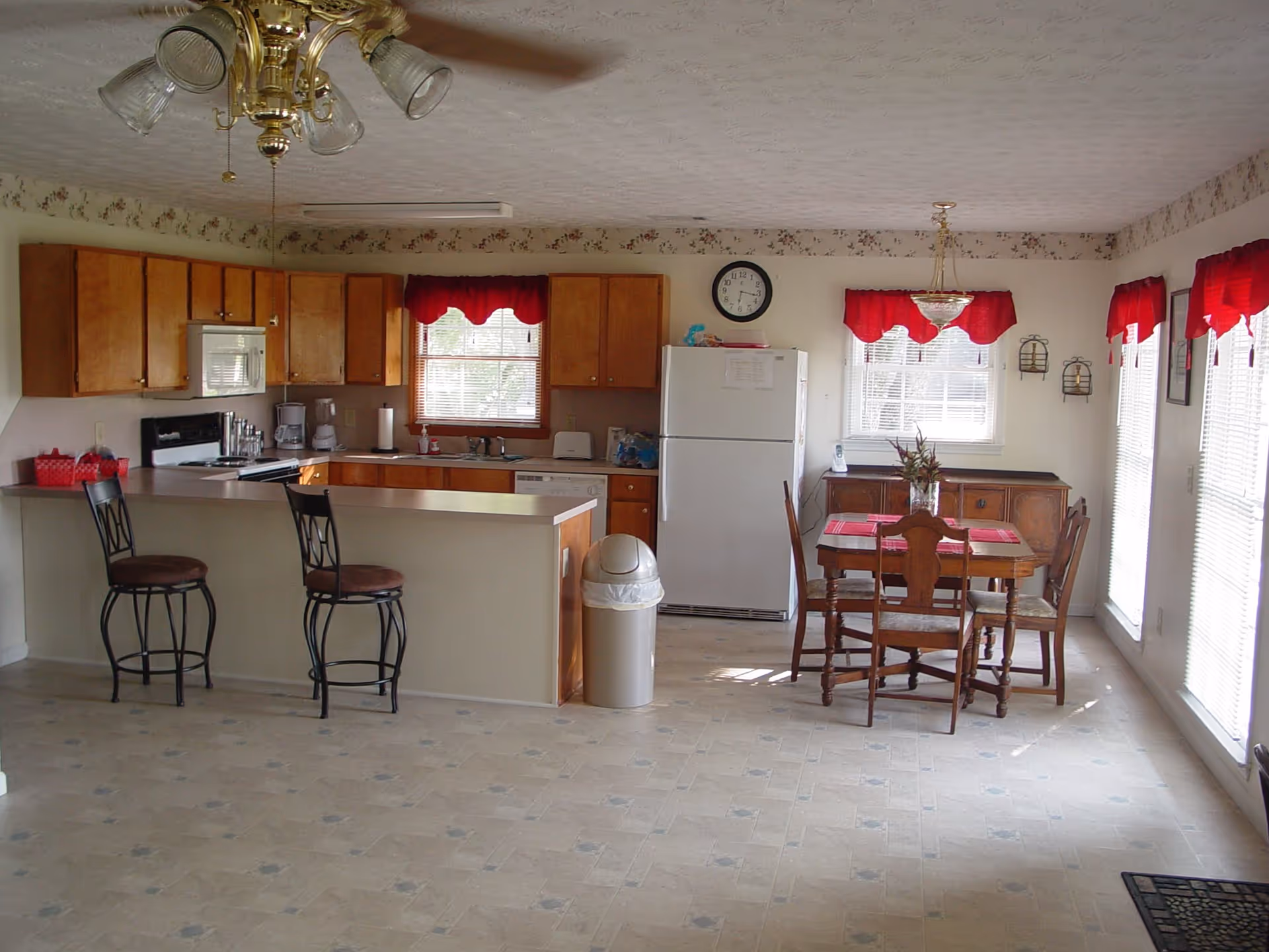 Open kitchen and dining area with a breakfast bar and two stools, wooden cabinets, refrigerator and a small dining table under red window valances.