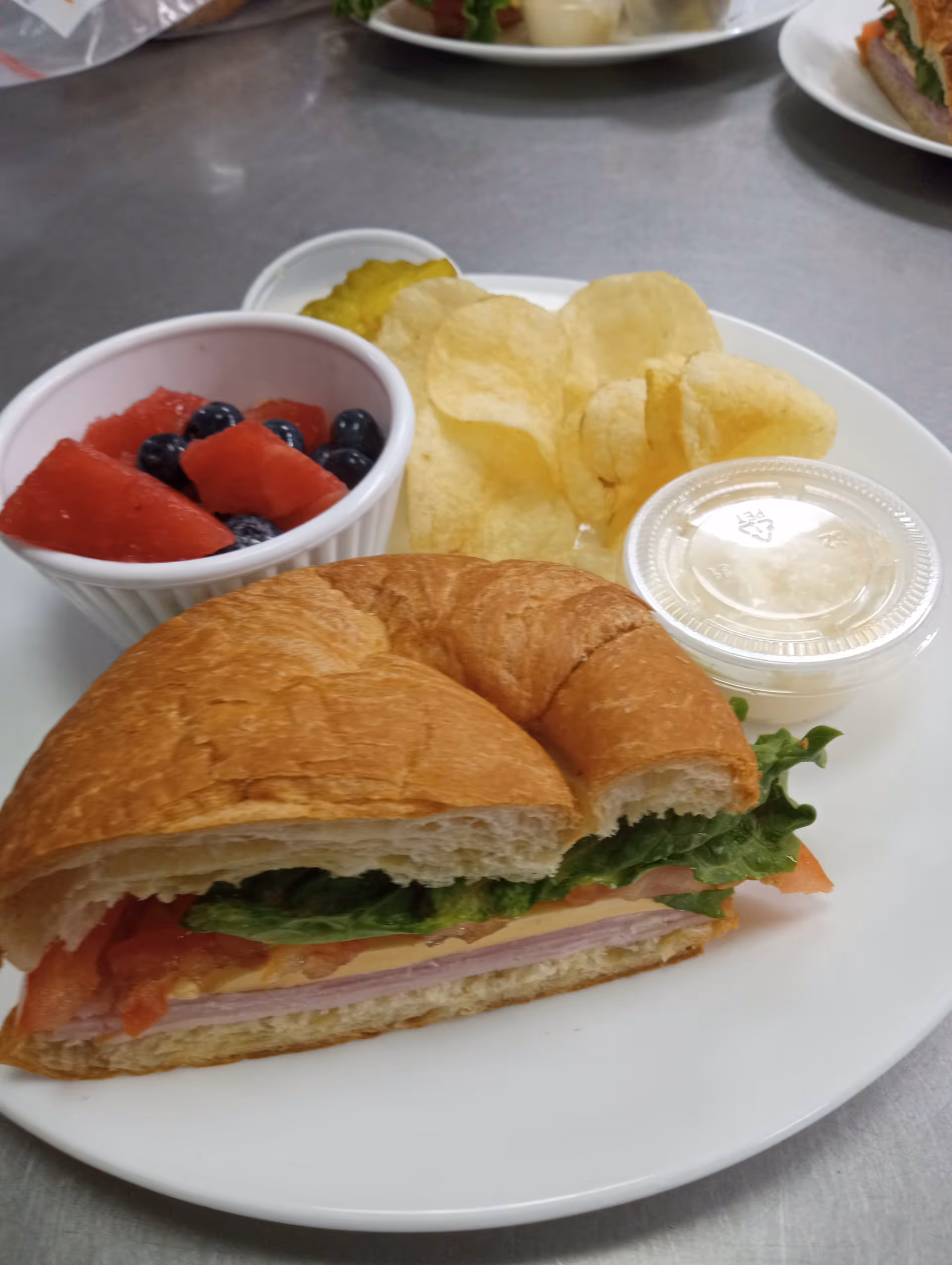 A plate with a croissant sandwich containing lettuce, tomato, cheese, and ham, accompanied by potato chips, a small container of pickles, a small cup of fruit salad with watermelon and blueberries, and a small container of dressing or sauce.