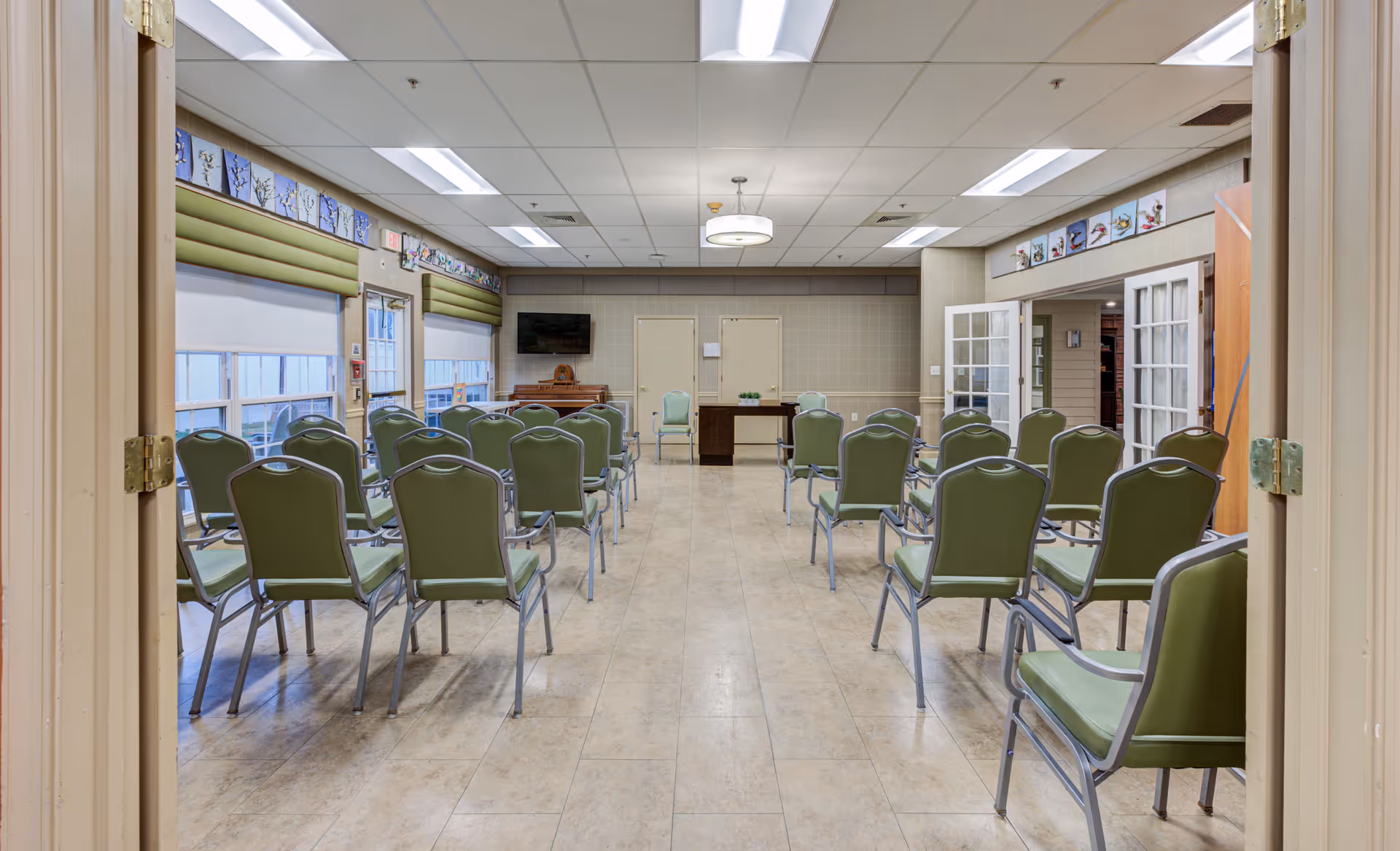 A meeting or activity room with rows of green chairs arranged facing a wall with two closed doors, a small desk, and a television mounted above a piano. The room has large windows on the left side with green valances and decorative artwork above the windows and doors. The ceiling has fluorescent lighting panels.