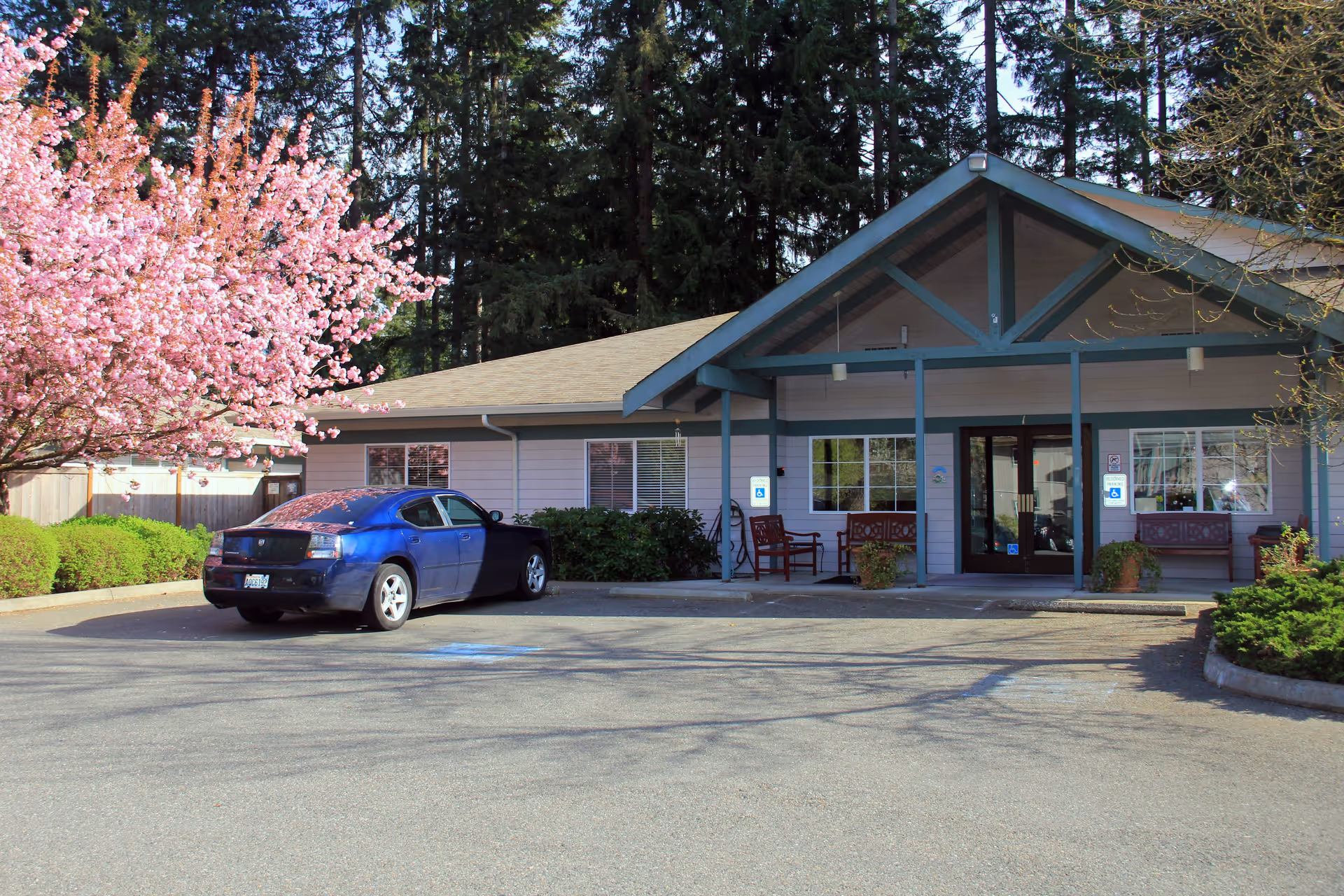 Exterior view of a single-story building with a covered entrance supported by blue beams. There are benches and potted plants near the entrance. A blue car is parked in a handicapped parking space in front of the building. To the left, there is a tree with pink blossoms and green shrubs. Tall evergreen trees are visible in the background.