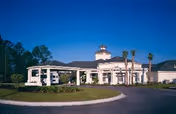 Exterior view of Mathison Retirement Community building at dusk with a circular driveway, palm trees, and well-maintained landscaping under a clear blue sky.