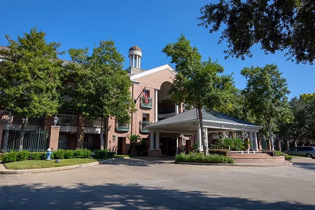 Exterior view of a multi-story brick building with a covered entrance, surrounded by trees and greenery under a clear blue sky.