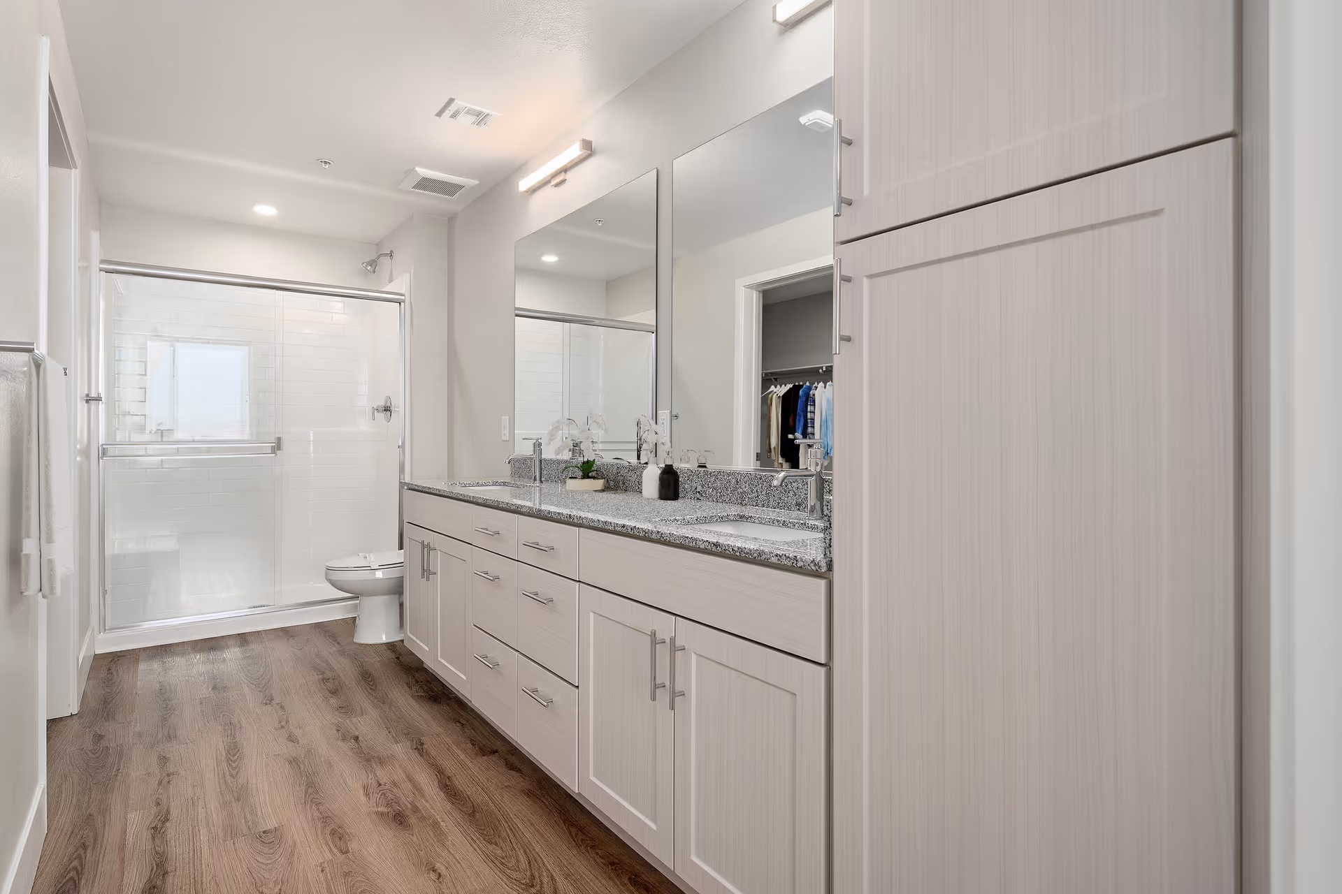 A modern bathroom with a double sink vanity featuring granite countertops and light wood cabinets. There is a large mirror above the sinks and a glass-enclosed shower with white subway tiles in the background. The floor is wood-style, and the room is well-lit with recessed lighting and a wall-mounted light fixture above the mirror.