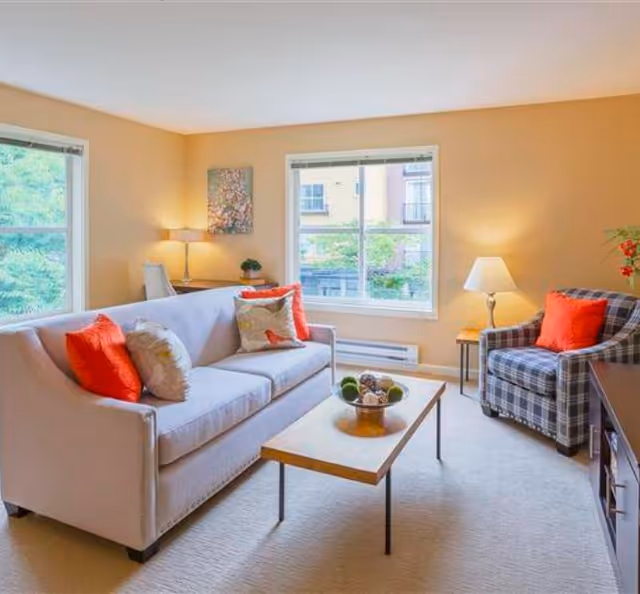 A bright living room with beige walls and carpet, featuring a light gray sofa with red and patterned throw pillows, a plaid armchair with a red pillow, a wooden coffee table with decorative items, two windows letting in natural light, a side table with a lamp, and a small desk with a chair and lamp in the corner.