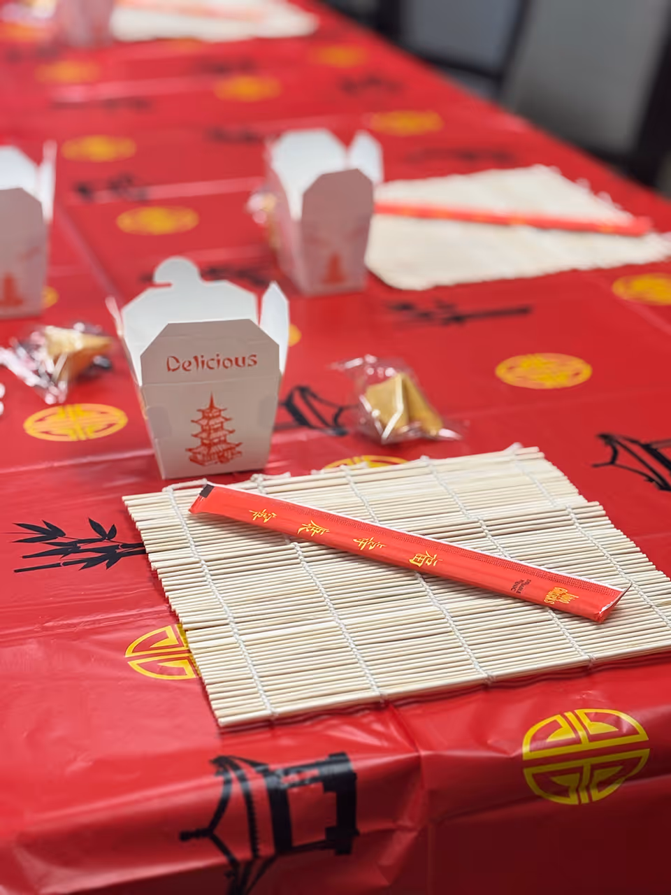 A red-decorated table with a bamboo mat, a red chopstick sleeve, a white takeout box labeled "Delicious", and fortune cookies.