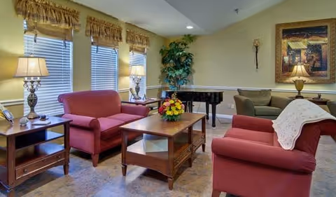 A cozy living room with two red upholstered armchairs and a matching loveseat arranged around a wooden coffee table with a flower arrangement. There are two side tables with lamps, a large potted plant in the corner, a black grand piano, and a framed painting on the wall. The room has three windows with blinds and valances, and the walls are painted a soft yellow.