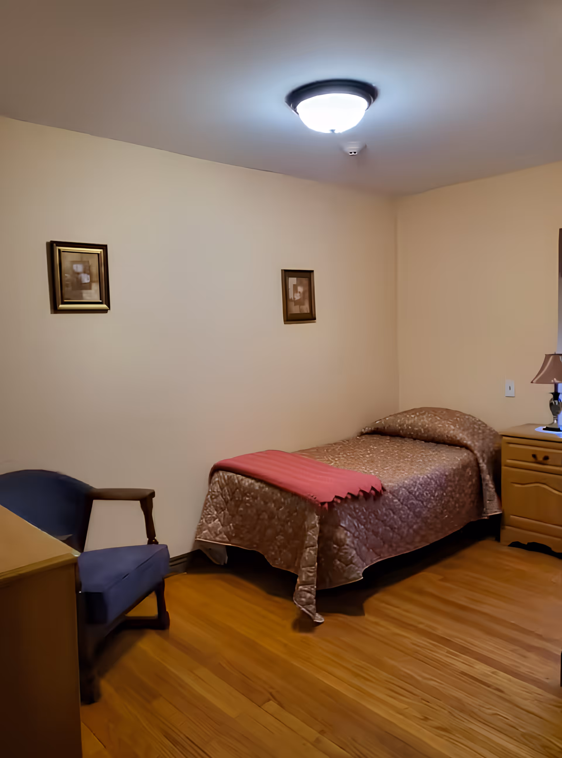 A small furnished bedroom with a single bed covered by a quilt and pink throw, a wooden nightstand with lamp, a chair, framed pictures on the wall, and hardwood floors.
