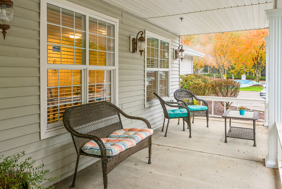 Covered front porch with wicker bench and chairs with colorful cushions, a small table, and autumn trees visible beyond the driveway.