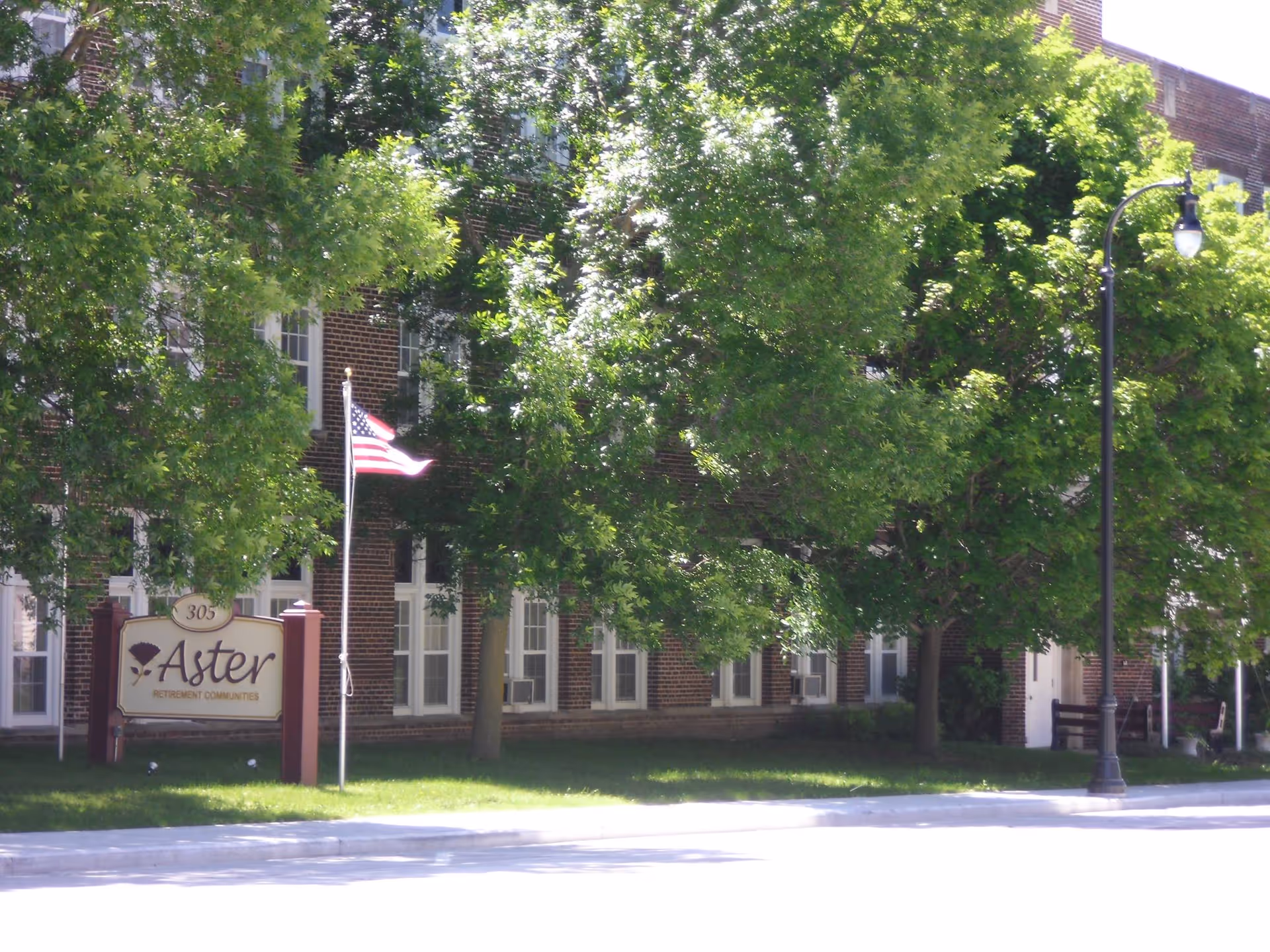 Front exterior of Aster Assisted Living brick building partially obscured by trees, with an American flag and a lawn sign that reads "Aster".