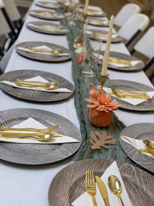 Long dining table set with wood-pattern chargers, gold flatware, white napkins, candles and fall-themed centerpieces.