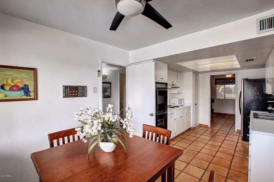 Dining area with a wooden table and white floral centerpiece next to a white kitchen with terracotta tile flooring and a ceiling fan.