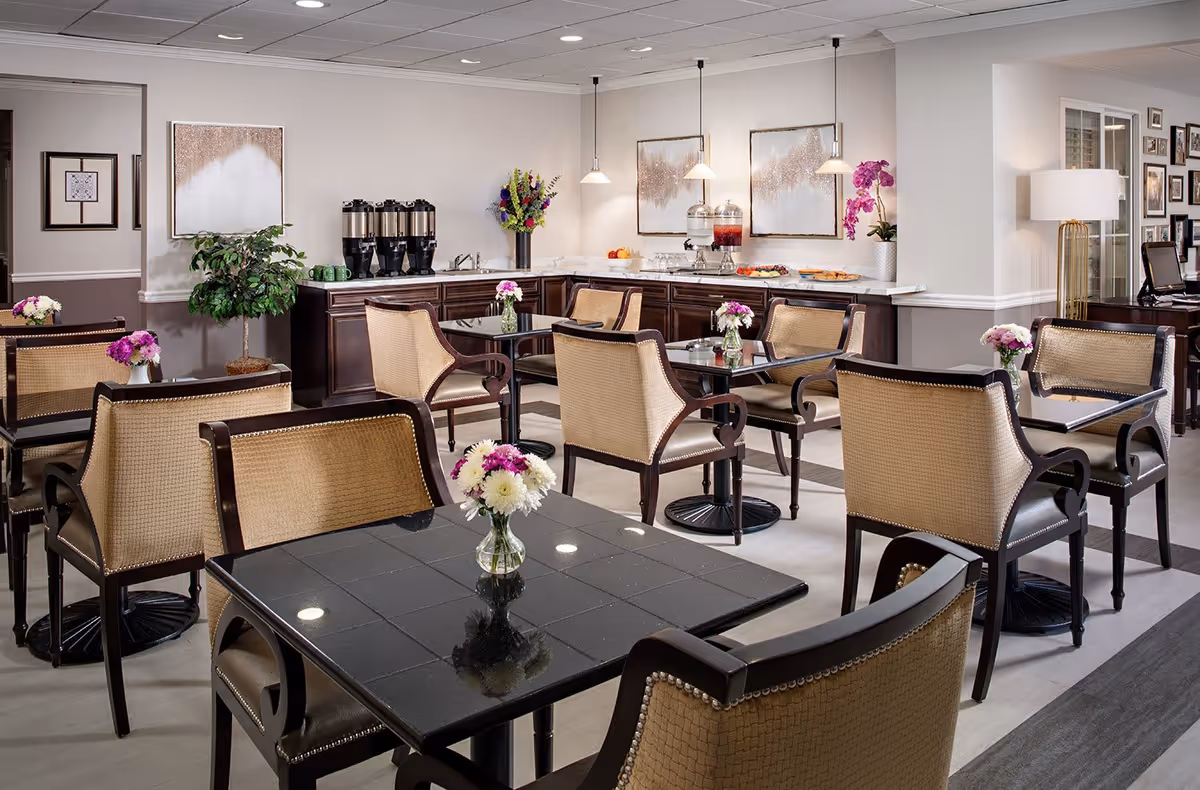 A well-lit dining area with several tables and cushioned chairs arranged neatly. Each table has a small vase with fresh flowers. In the background, there is a counter with beverage dispensers, cups, and plates of food. The walls are decorated with framed artwork and a potted plant is placed near the counter.