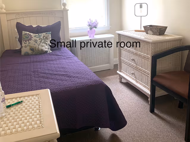 Small private bedroom with a single bed dressed in a purple quilt, wicker dresser, chair, and a window with flowers.