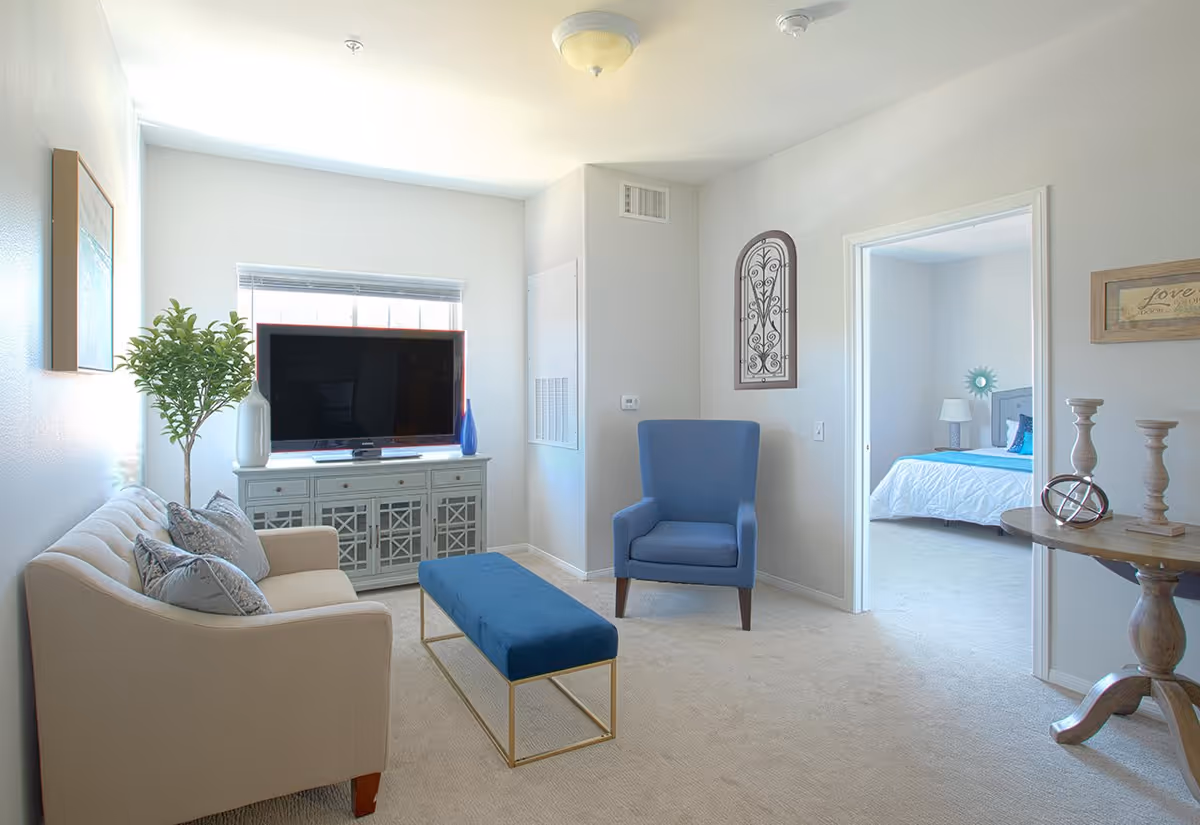 A bright living room with a beige sofa adorned with pillows on the left, a blue upholstered armchair on the right, and a blue cushioned bench with gold legs in the center. A TV sits on a decorative gray cabinet against the far wall beneath a window with blinds. A potted plant is next to the sofa, and a round wooden side table with decorative items is on the right. An open doorway leads to a bedroom with a bed, nightstand, and lamp.