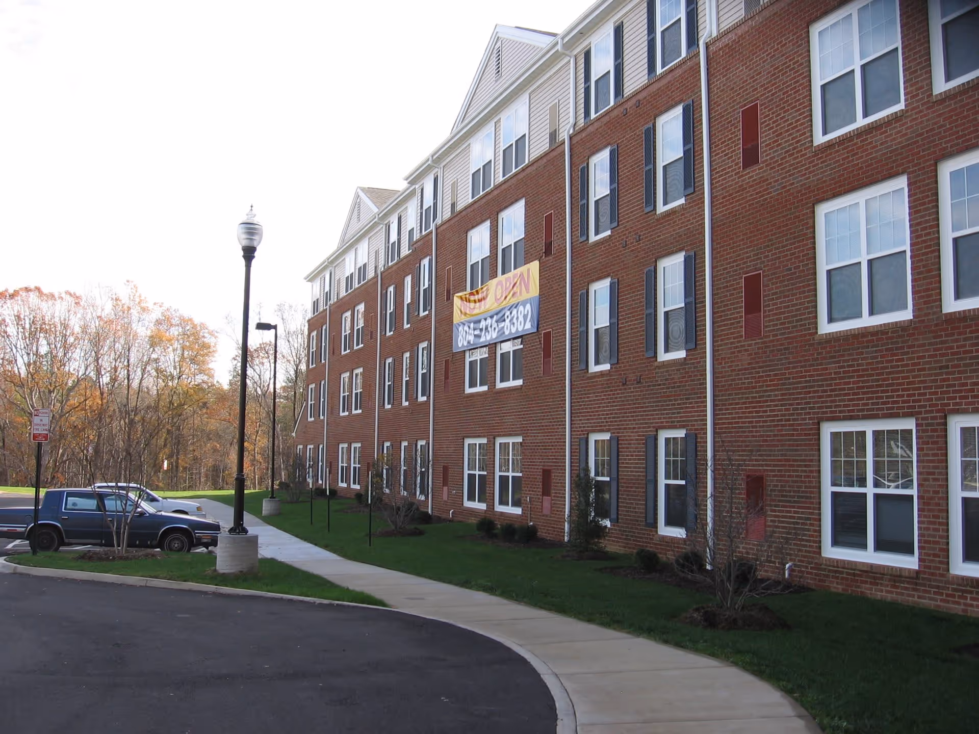 Exterior view of a multi-story brick building with white-framed windows and black shutters. A sidewalk curves along the front of the building, with a streetlamp and parked vehicles visible. Trees with autumn foliage are in the background. A banner on the building reads 'NOW OPEN' with a phone number.