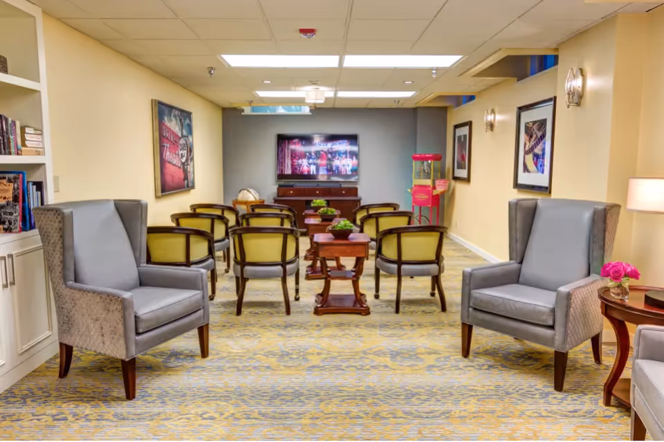 A cozy common area with several chairs arranged in rows facing a wall-mounted television. The room has a patterned carpet, soft yellow walls, framed artwork, a popcorn machine in the corner, and a couple of side tables with decorative plants and flowers.