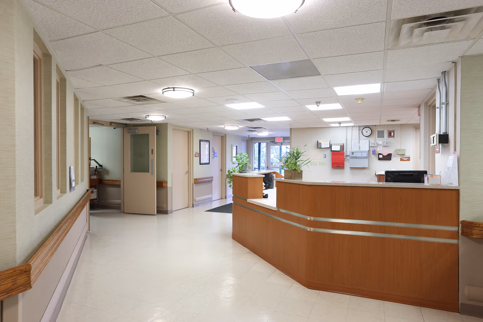 A clean and well-lit reception area in a senior living facility with a wooden front desk, beige walls, ceiling lights, and plants near the entrance door in the background.
