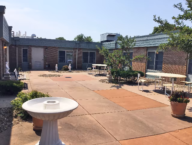 Outdoor courtyard area with paved walkways, a white birdbath in the foreground, potted plants, small trees, and several tables and chairs. The courtyard is surrounded by a single-story brick building with windows and doors.