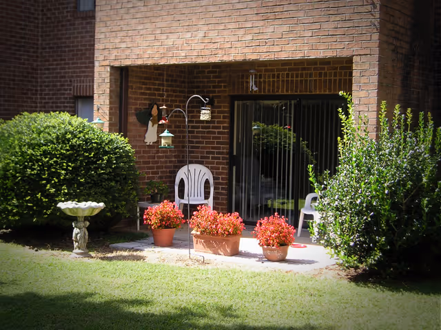 A ground-level brick patio with a sliding glass door, two white plastic chairs, potted red flowers, a birdbath, and surrounding shrubs.
