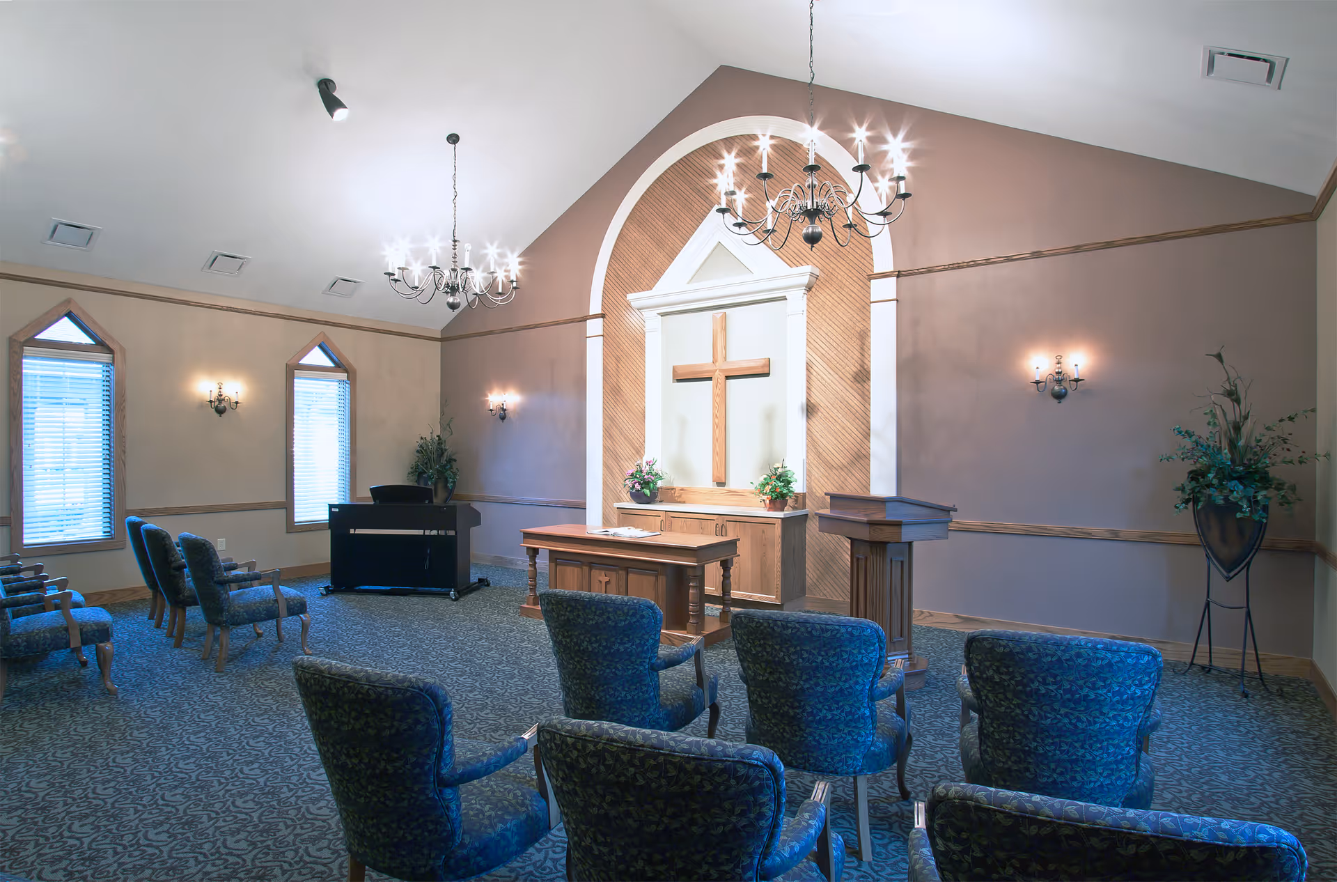 Interior view of a chapel or small worship room with a wooden cross mounted on a white panel at the front. The room has several upholstered chairs arranged in rows facing the cross, two chandeliers hanging from the ceiling, wall sconces with lights, two tall windows with blinds, a piano, and floral arrangements on either side of the cross.