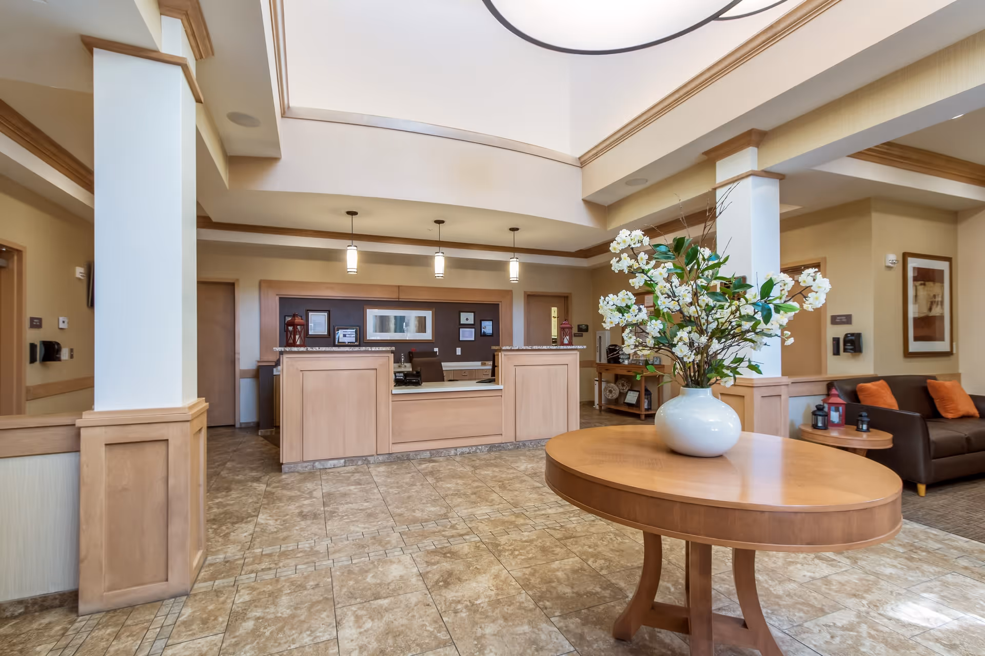 A bright and spacious reception area with a wooden front desk, beige tiled floor, and a round wooden table with a white vase holding white flowers. The area has beige walls, a high ceiling with recessed lighting, and a seating area with a brown couch and orange cushions to the right.