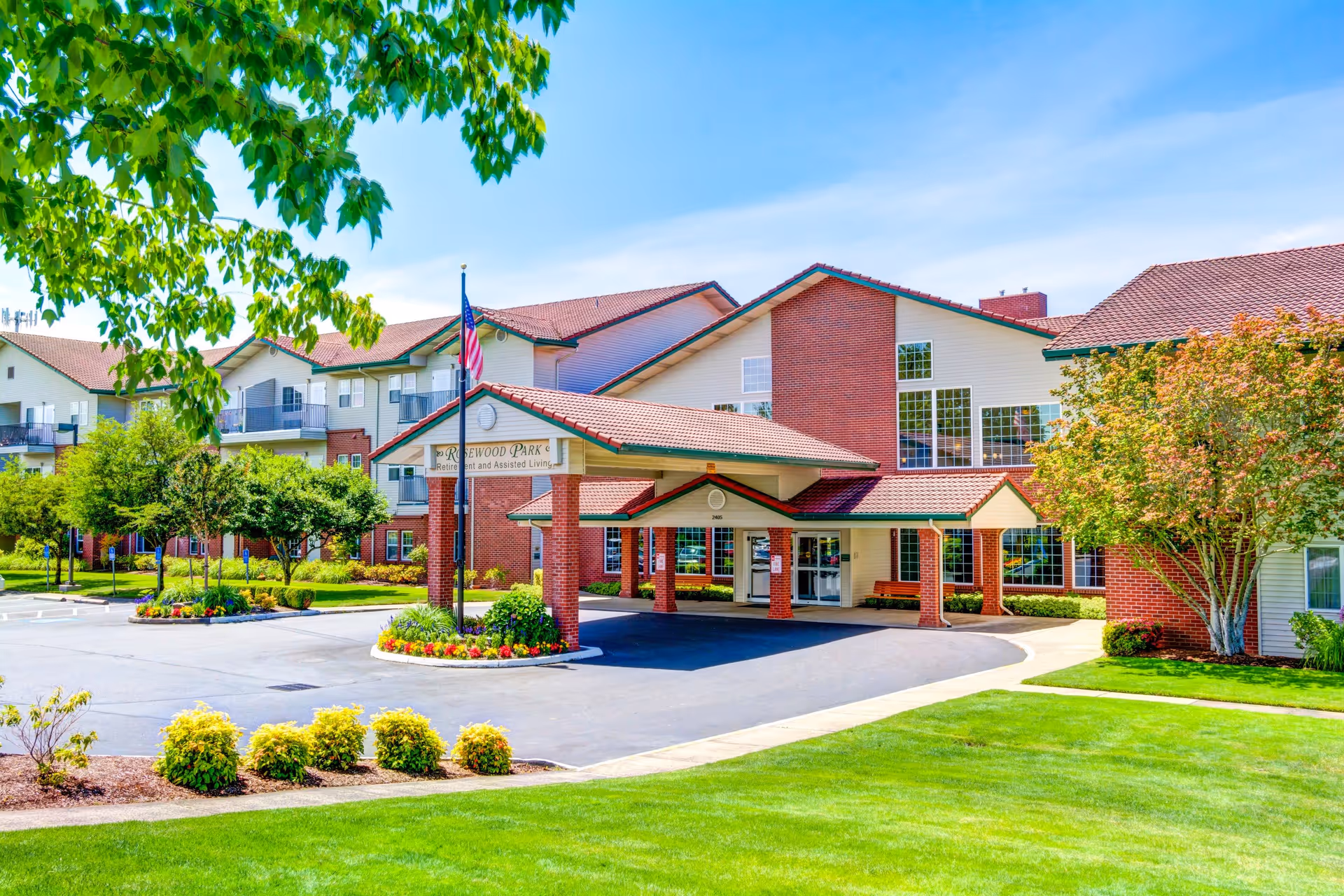 Front entrance of the Rosewood Park senior living facility showing the covered porte-cochère, American flag, and landscaped grounds.