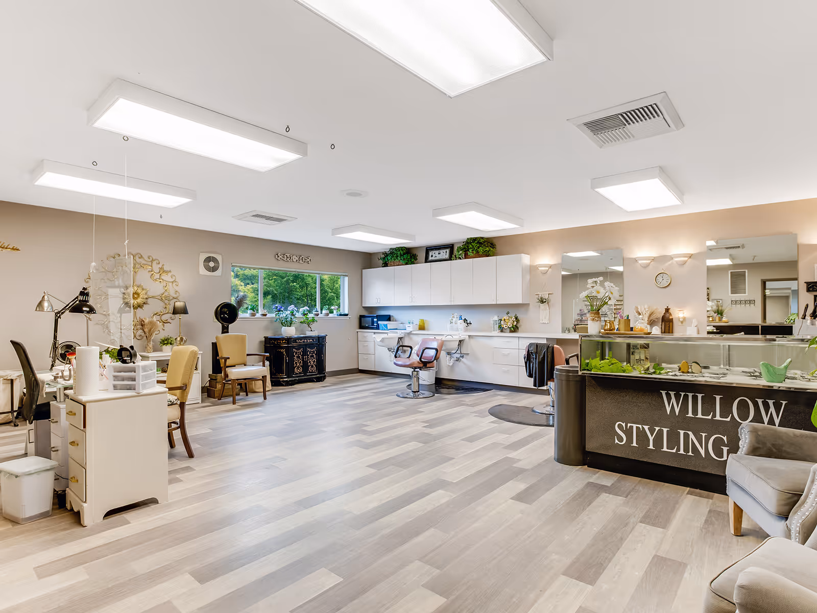 A spacious and well-lit hair styling salon area with multiple styling chairs, mirrors, and cabinets. The room has light wood flooring, large overhead lights, and a window with a view of greenery outside. There are decorative plants and flowers on the counters and shelves, and a sign on the front desk reads 'Willow Styling'.