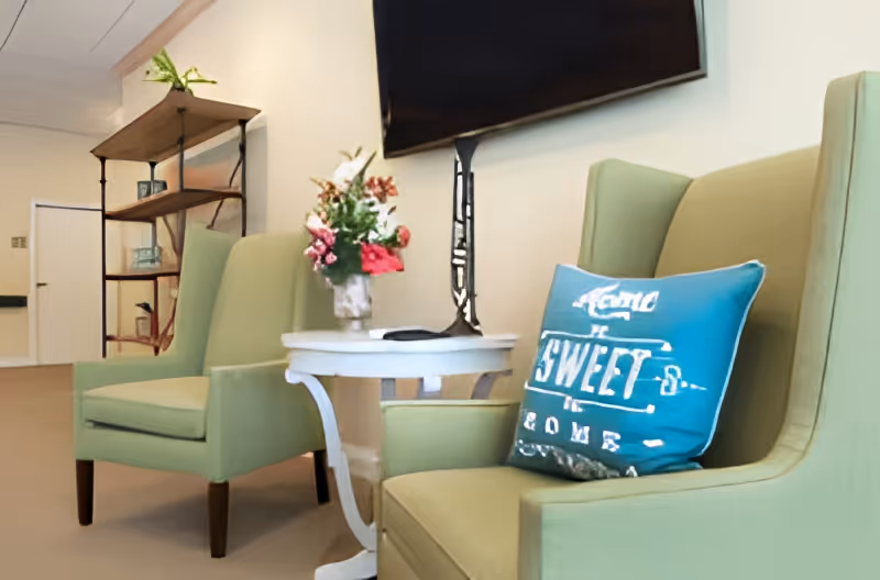 A cozy seating area with two light green armchairs, a small white round table between them holding a vase with flowers, and a blue decorative pillow on one chair that says 'Home Sweet Home'. A flat-screen TV is mounted on the wall above the table, and a wooden shelving unit with plants and decorative items is visible in the background.
