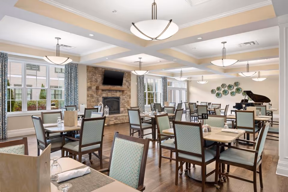 A spacious dining room with multiple tables and chairs arranged neatly. The room features large windows with blue patterned curtains, a stone fireplace with a mounted flat-screen TV above it, and a grand piano in the corner. The ceiling has recessed lighting and modern hanging light fixtures. Tables are set with placemats, napkins, and glasses.