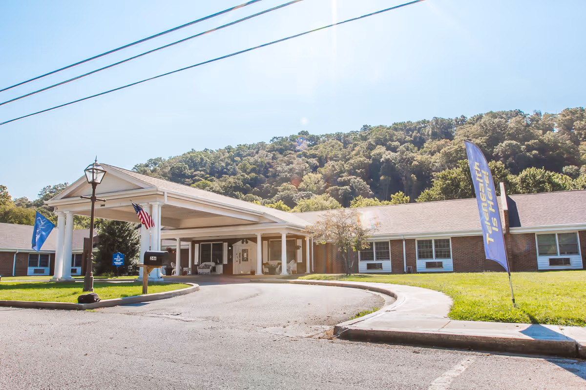 Exterior view of Lee County Care and Rehabilitation Center showing a single-story brick building with a covered entrance supported by white columns. There are flags near the entrance, including an American flag and a blue flag with text. The building is surrounded by green grass and trees, with a wooded hill in the background under a clear sky.
