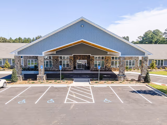Front entrance of a single-story assisted living facility with a large covered porte-cochère, stone columns, and marked handicap parking spaces in front.