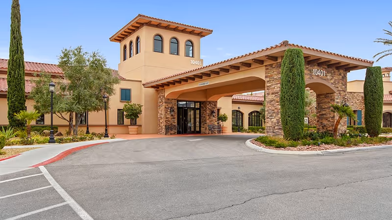 Exterior view of a senior living facility named Las Ventanas at Summerlin, featuring a beige and stone facade with a covered entrance, tall windows, and landscaped greenery including trees and shrubs.