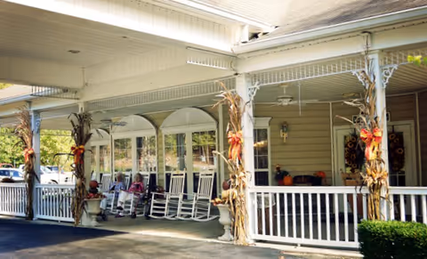 Covered front porch with white rocking chairs, fall-decorated columns, and seated residents at the assisted living entrance.